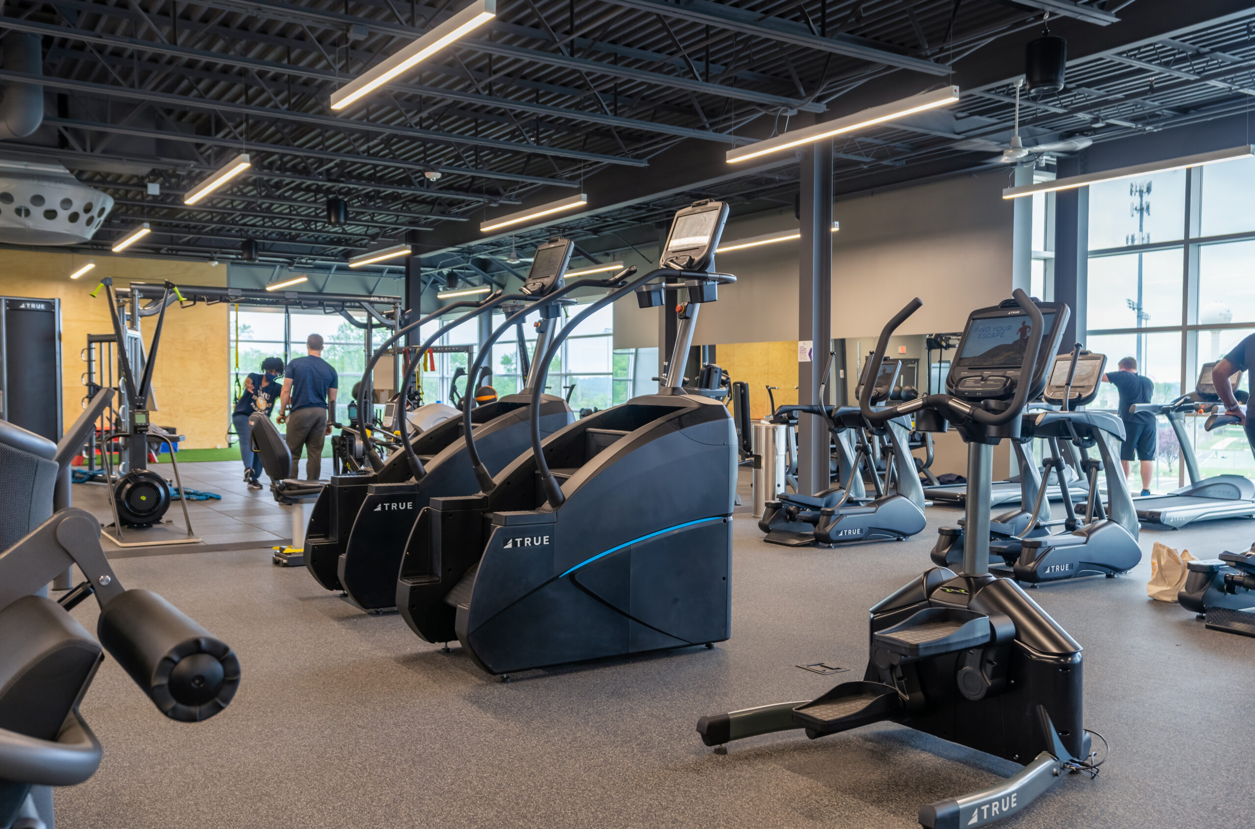 A modern gym with various exercise machines, including stair steppers, stationary bikes, and ellipticals. A few people are working out, and large windows let in natural light. The space has an open, industrial feel.