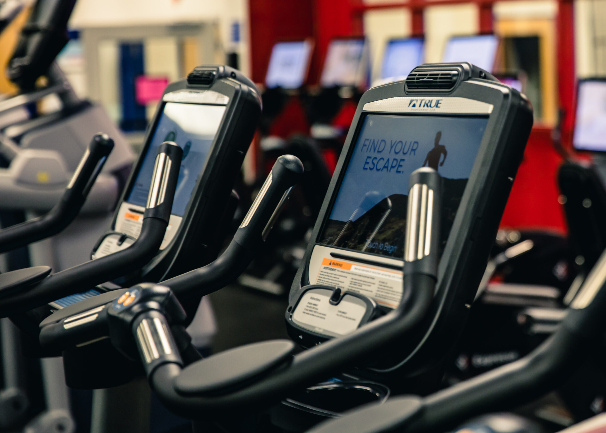 Close-up of exercise bikes with digital screens in a gym. The nearest screen displays a message, Find your escape, with a mountain image. Other gym equipment and screens are visible in the blurred background.