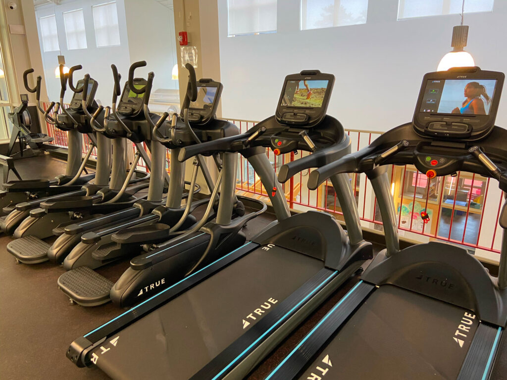 A row of treadmills with digital screens stands in a well-lit gym, next to a railing overlooking a lower floor with colorful play equipment and windows letting in natural light.