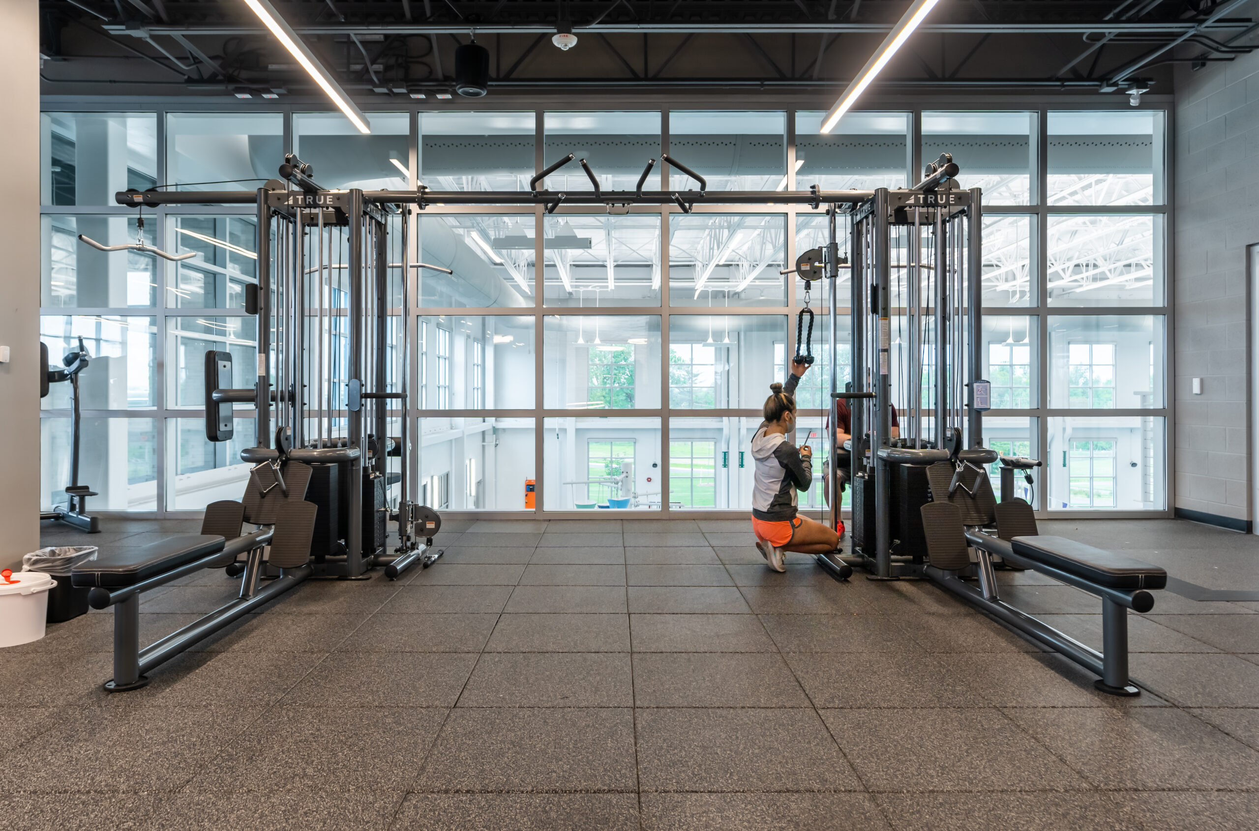 A person in athletic clothes kneels while using a cable machine in a spacious, modern gym with large windows and abundant natural light. The gym appears clean and empty except for the individual.