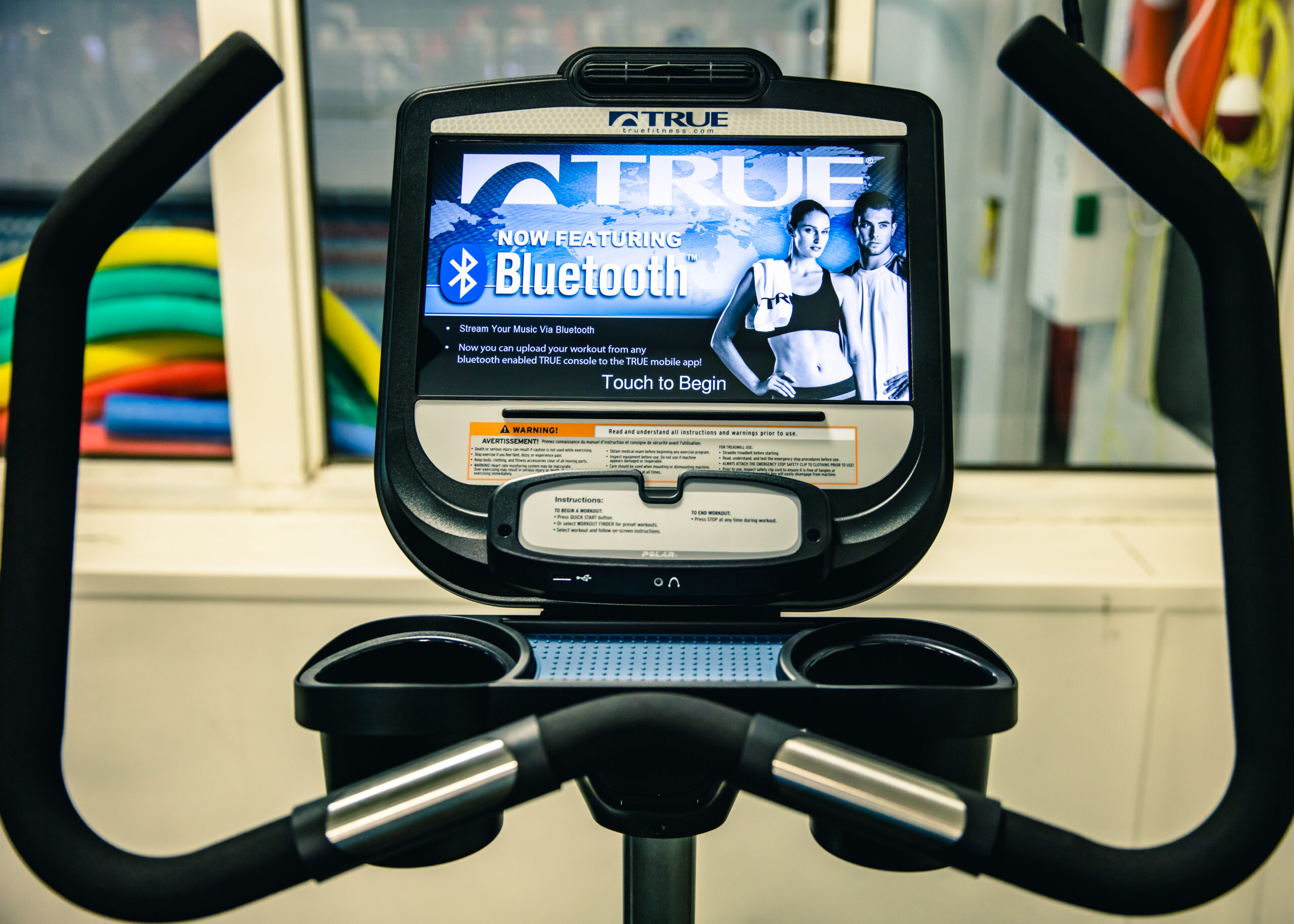 A close-up of a treadmill display screen showing a Bluetooth feature advertisement, with two cup holders and handlebars in the foreground; colorful exercise equipment is visible through a window behind.