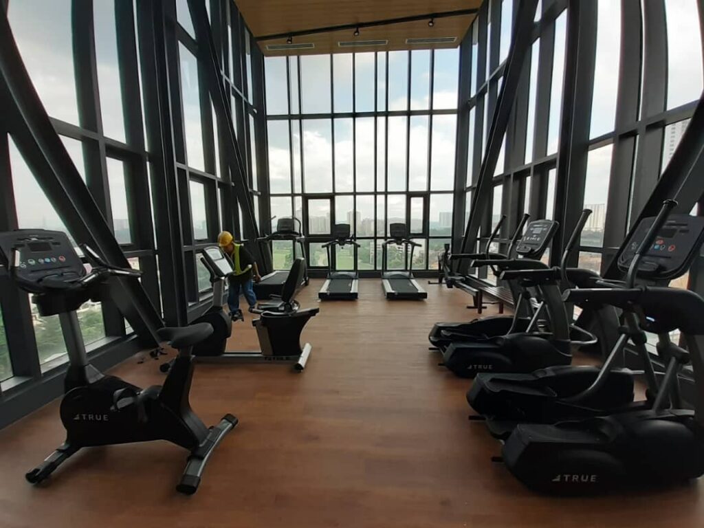 A modern gym with large windows, filled with exercise bikes and treadmills on a wooden floor, offering a city view through the glass walls.