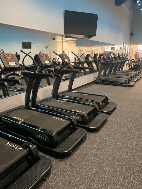 A row of treadmills lined up in a gym, facing a large wall mirror. Several have screens, and a large TV is mounted on the wall above them. The room has gray flooring and bright lighting.