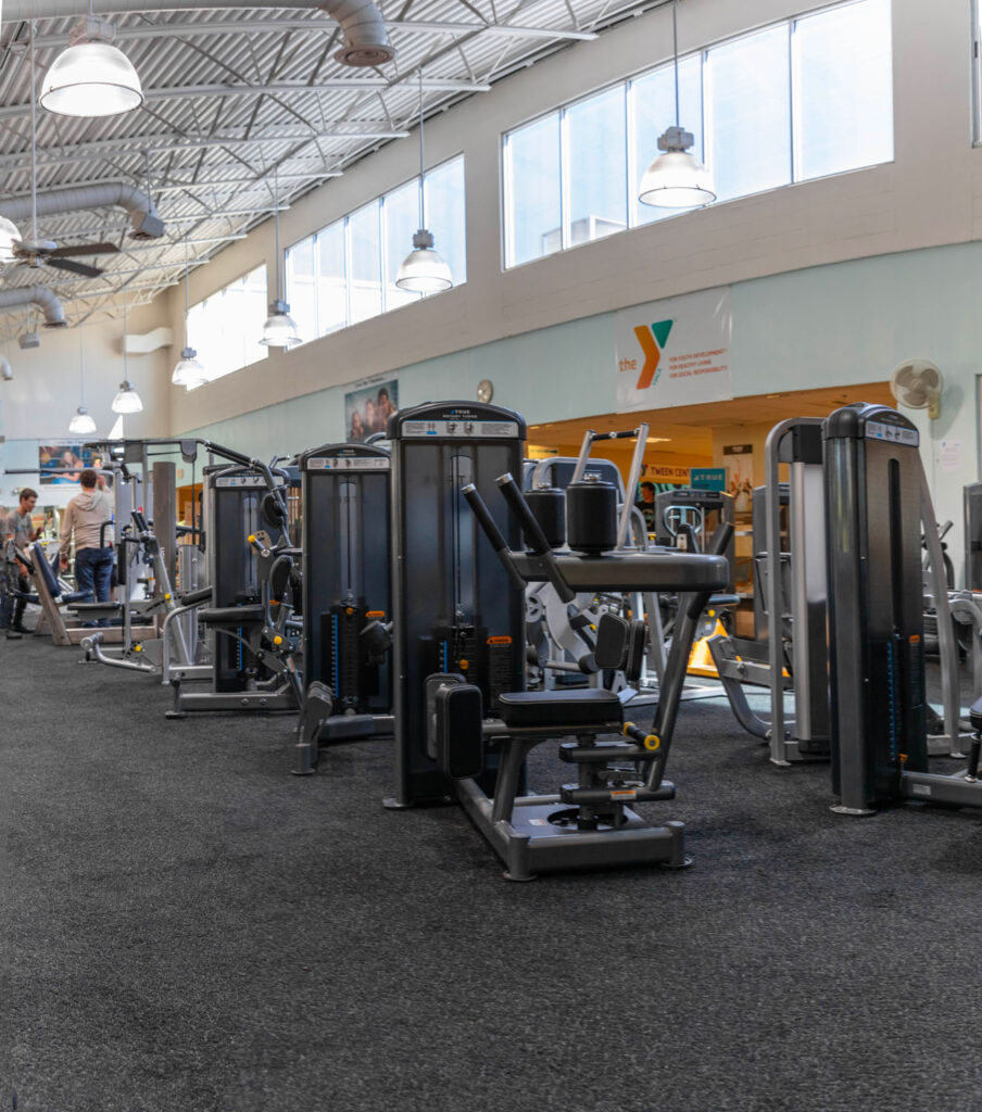 A spacious gym with various exercise machines on a black rubber floor, large windows letting in natural light, and a YMCA logo on the wall in the background. Several people are walking near the entrance.