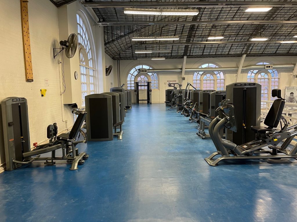 A spacious gym with blue floors, large arched windows, and several weight and resistance machines lined up along the walls. The gym appears empty and well-lit.