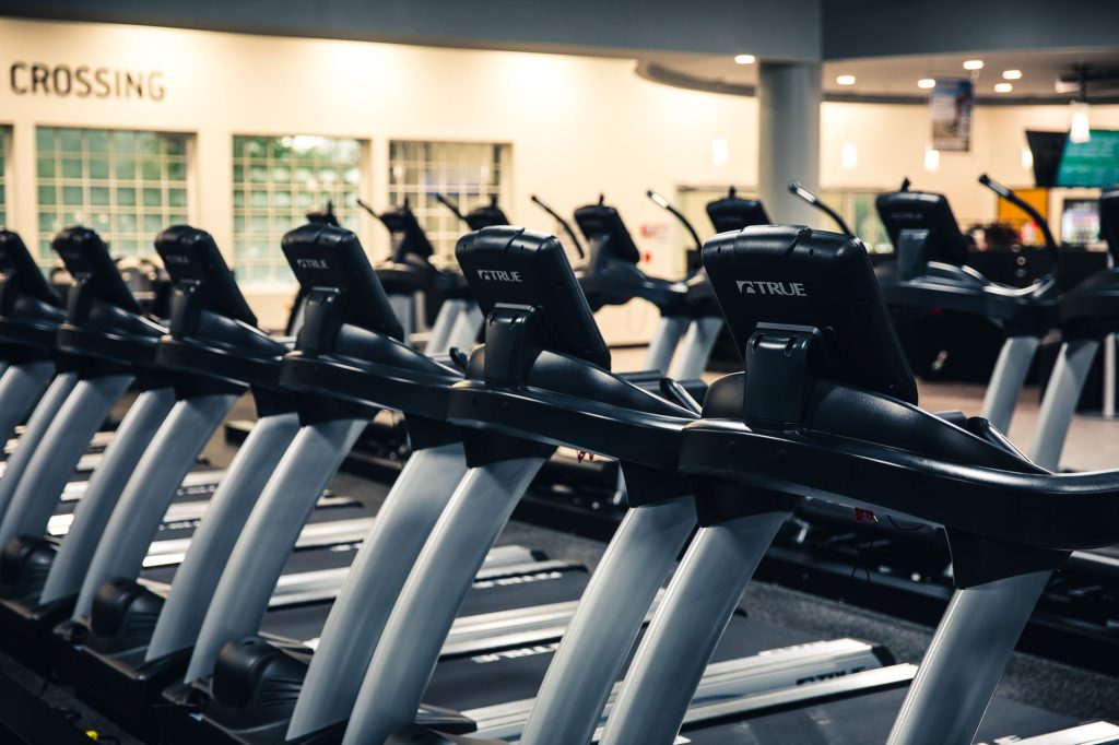 Rows of black treadmills are lined up in a well-lit, modern gym with large windows and a sign on the wall that says CROSSING in the background.