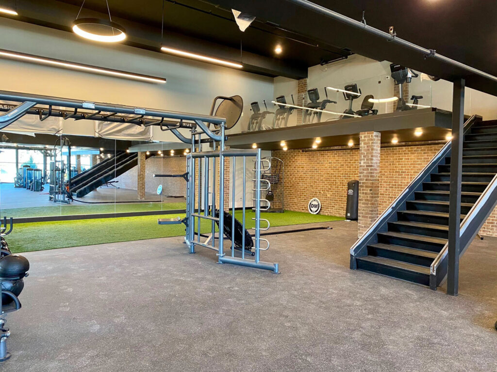 Modern gym interior with exercise equipment, free weights, and turf strip on the ground floor. A staircase leads to an upper level with cardio machines by glass railing and brick walls throughout. The space is brightly lit and spacious.