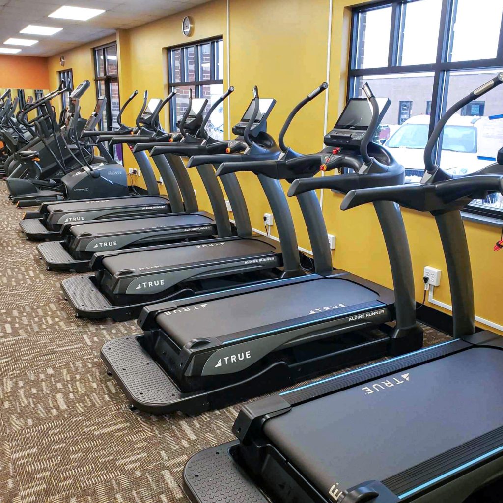A row of black treadmills lined up in a gym with yellow walls and large windows letting in natural light.