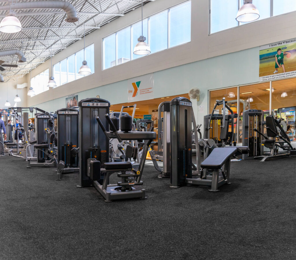 A spacious gym with various exercise machines on a black rubber floor, large windows along one wall, bright lighting, and a YMCA logo on the back wall.