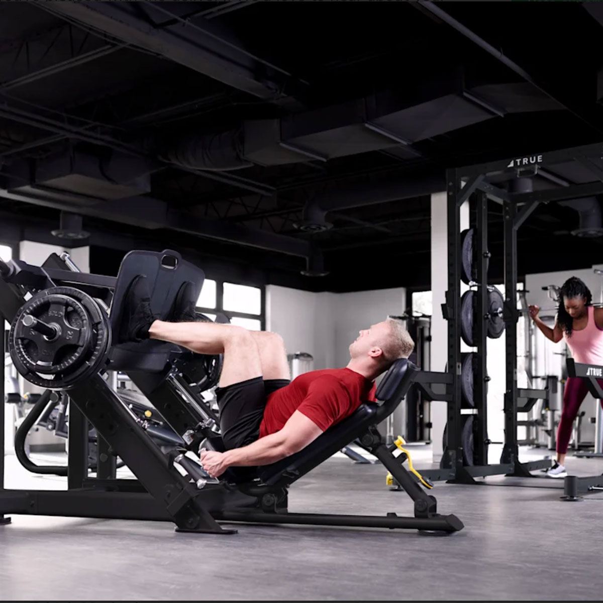 A man in a red shirt and black shorts uses a leg press machine in a gym, while a woman in athletic wear works out in the background near weight racks.