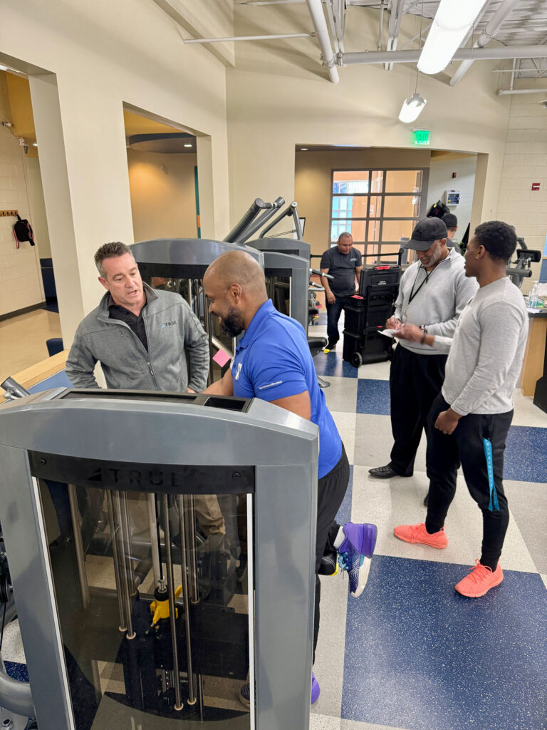 Four men stand and talk around gym equipment in a fitness center, while another man in workout clothes uses a machine. The space has bright lighting and exercise machines in the background.