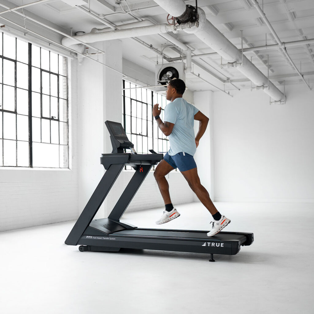 A person in athletic clothing runs on a treadmill in a bright, spacious room with large windows and exposed pipes on the ceiling. The room appears modern and minimalistic.