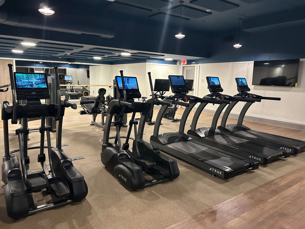 A modern gym with several treadmills and elliptical machines lined up, each equipped with screens. The room has bright lighting, mirrors on the walls, and a clean, organized appearance.