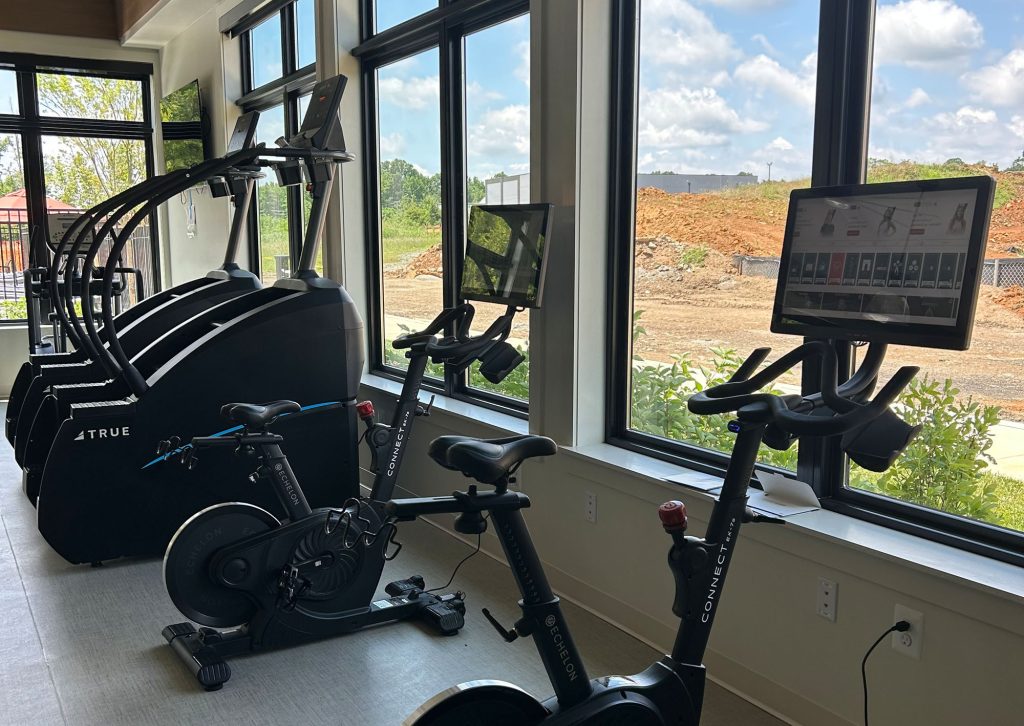 A bright gym with large windows features two stationary bikes and three stair climbers, each equipped with screens. Greenery and a construction site are visible outside under a partly cloudy sky.