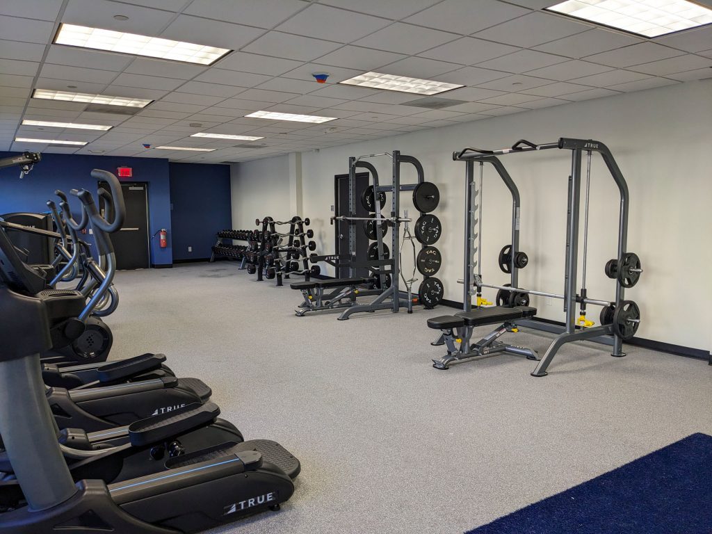 A modern gym with treadmills, ellipticals, free weights, weight benches, and weight racks on a carpeted floor under bright ceiling lights. The walls are white and blue, and theres a fire extinguisher in the back.