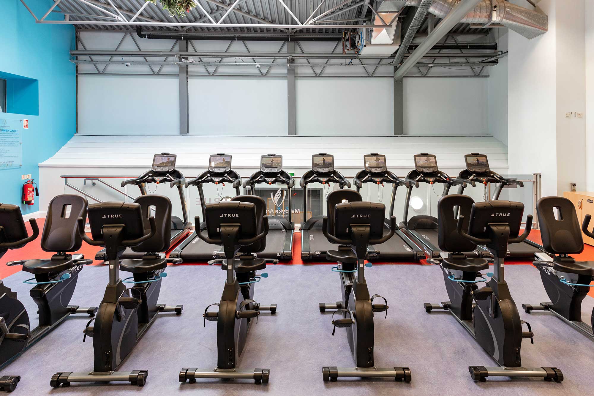 A row of stationary bikes and treadmills are lined up in a modern gym with high ceilings, exposed pipes, and bright lighting. The space appears clean and organized.