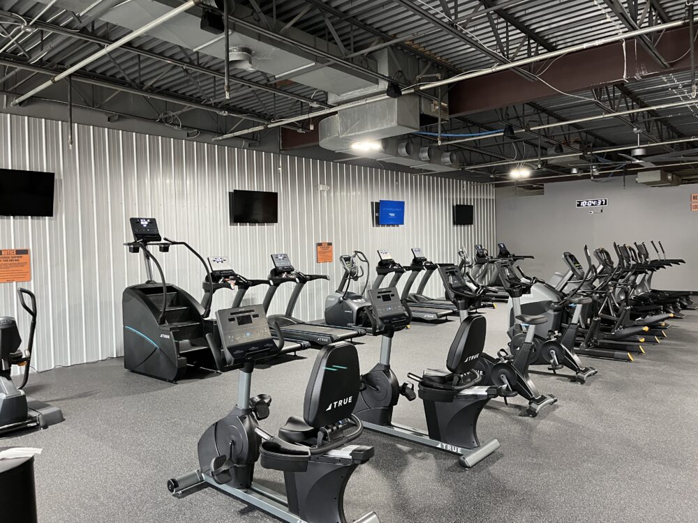 Rows of exercise machines, including stationary bikes, ellipticals, and treadmills, are arranged in a spacious gym with gray walls, multiple TVs mounted on the wall, and exposed ceiling beams.