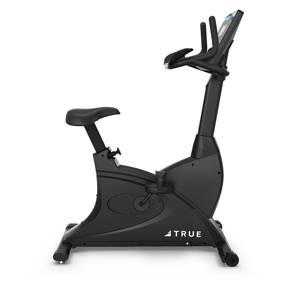 A black stationary exercise bike with a digital display, wide seat, and handlebars, positioned on a light background. The brand name TRUE is visible on the lower frame.