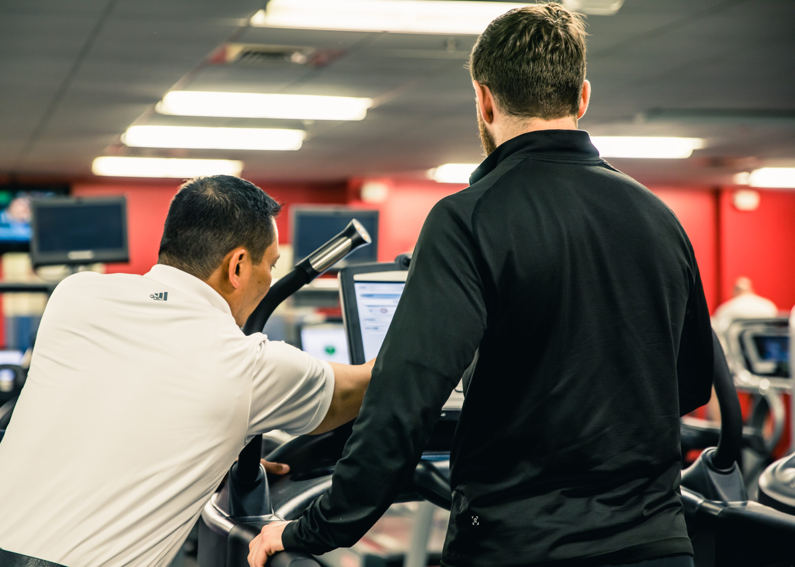 Two men in athletic clothing stand on treadmills in a gym; one points at the treadmill’s screen, appearing to explain or assist, while the other looks on. The gym has red walls and multiple exercise machines.