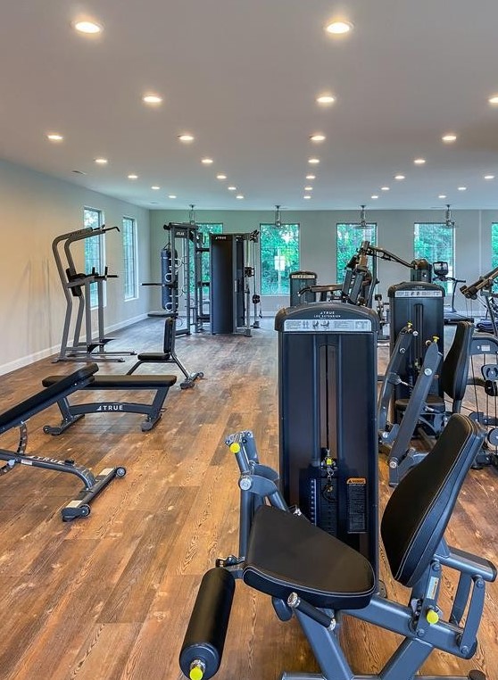 A spacious, well-lit gym with various exercise machines, benches, and large windows along the back wall letting in natural light. The room has wood floors and modern fitness equipment neatly arranged.