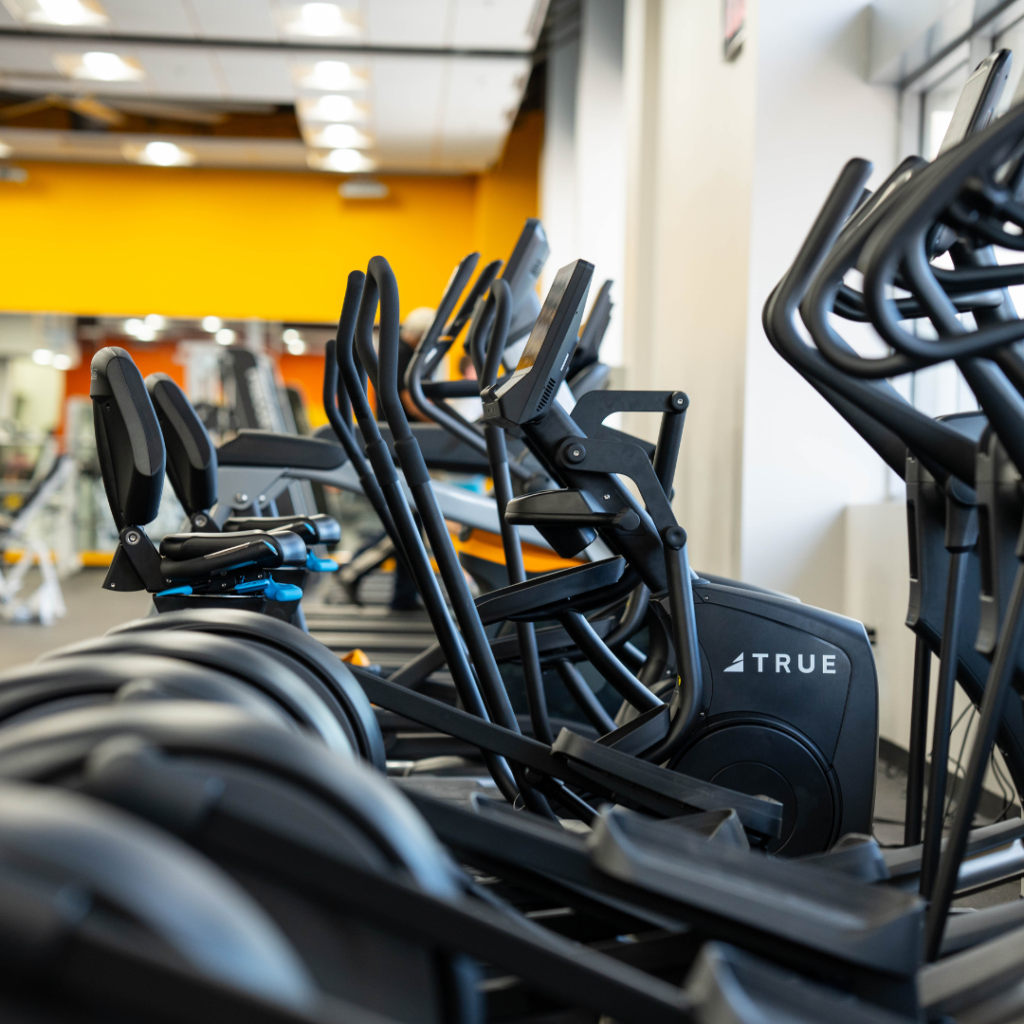 A row of elliptical machines in a brightly lit gym with yellow and white walls, viewed from the side, showing the equipment neatly lined up and ready for use.