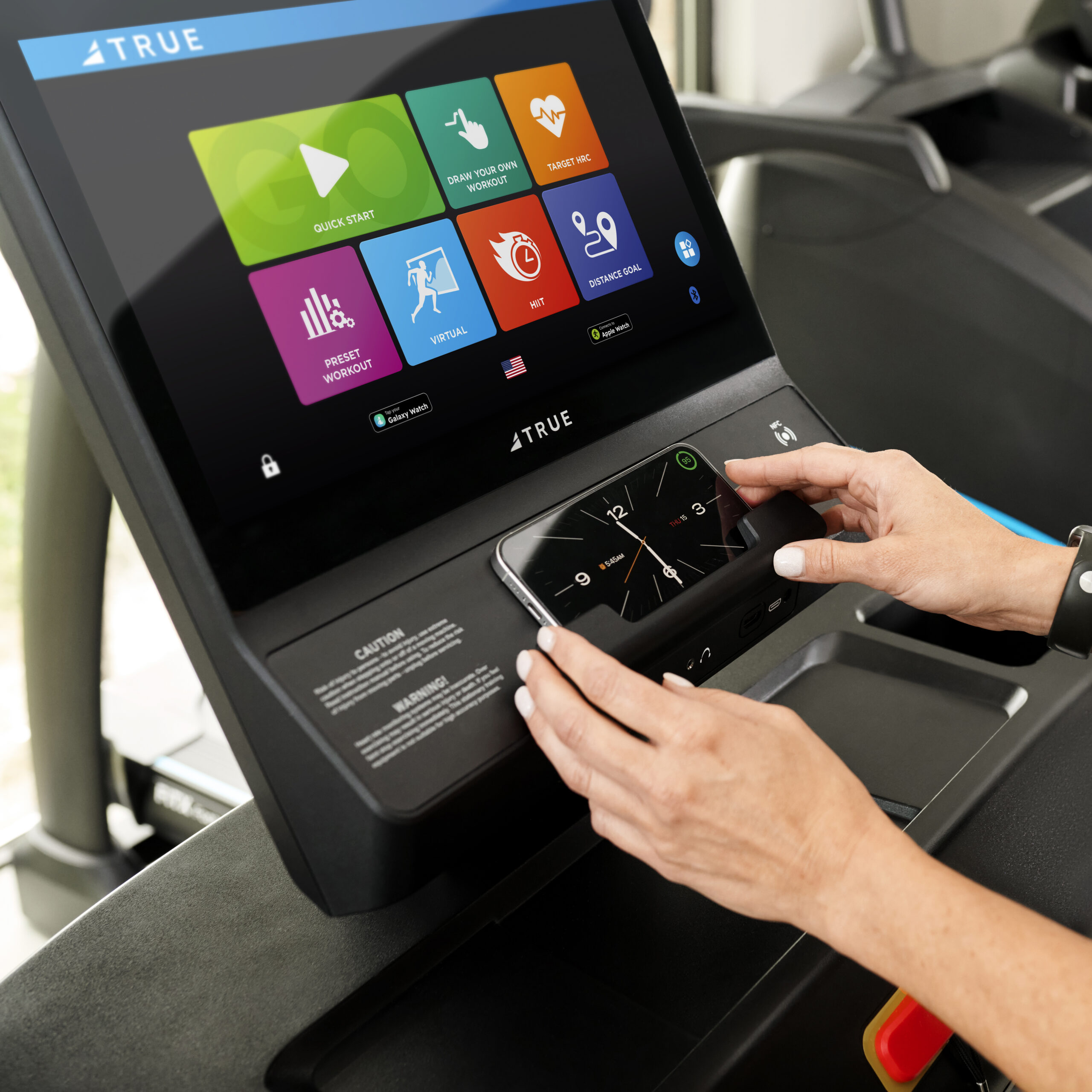A person places a smartphone on the console of a treadmill, which displays a touchscreen with workout options like Quick Start, Fitness Test, and TV.