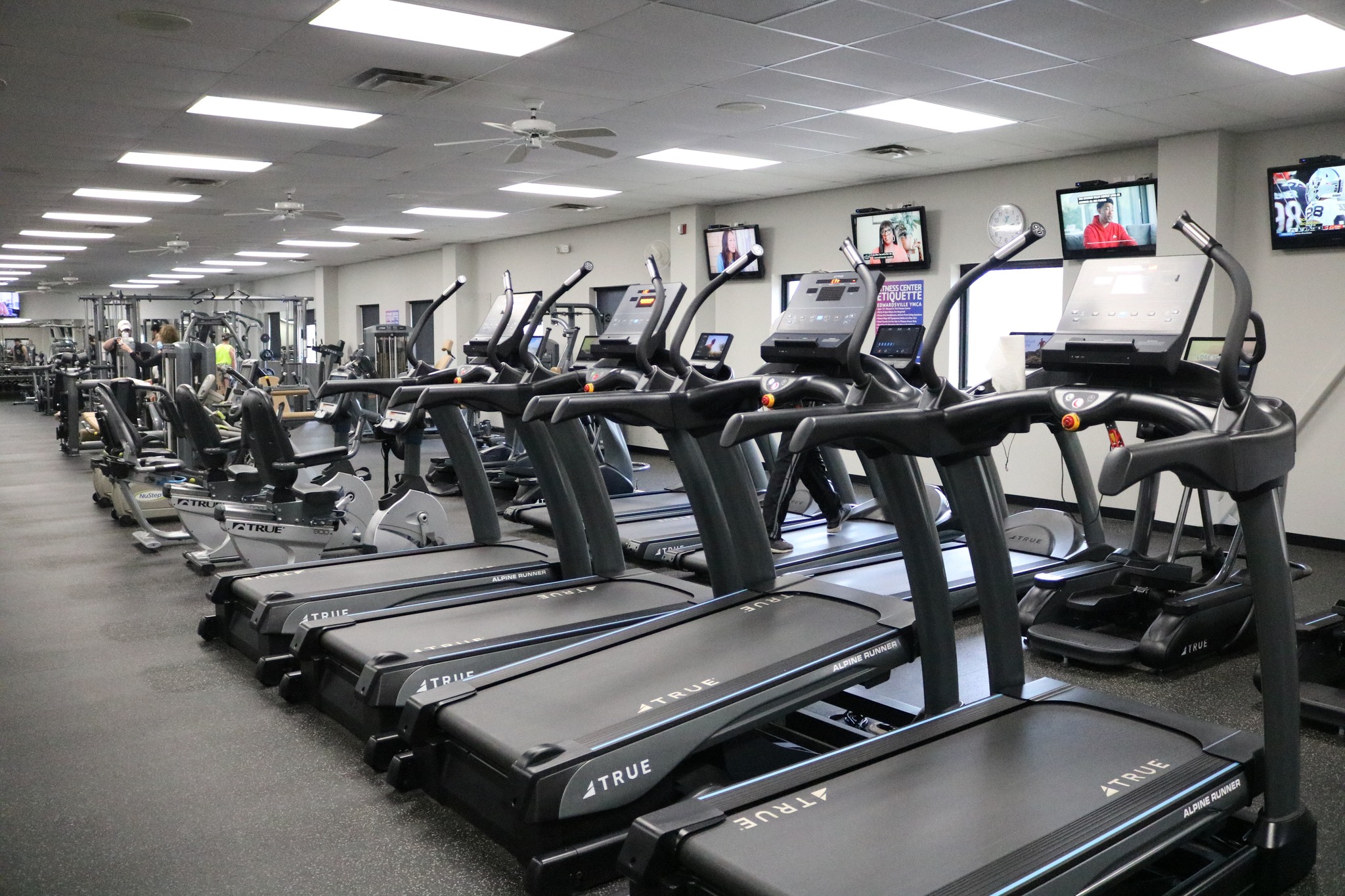 A spacious gym with rows of treadmills, exercise bikes, and other fitness equipment; several wall-mounted TVs are on, and a few people are working out in the background. The gym has bright lighting and a clean, organized layout.