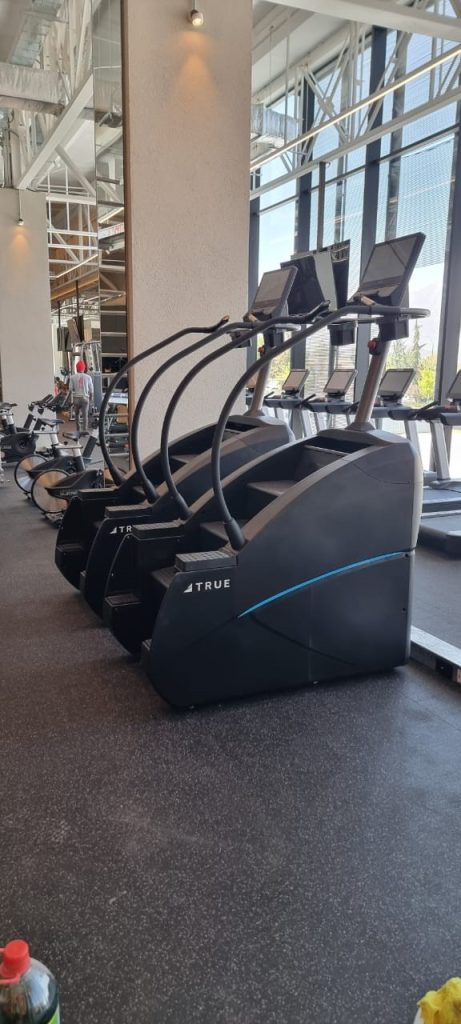 Three stair-climber machines are lined up in a modern gym with large windows and reflective surfaces. Other gym equipment and exercise bikes are visible in the background.