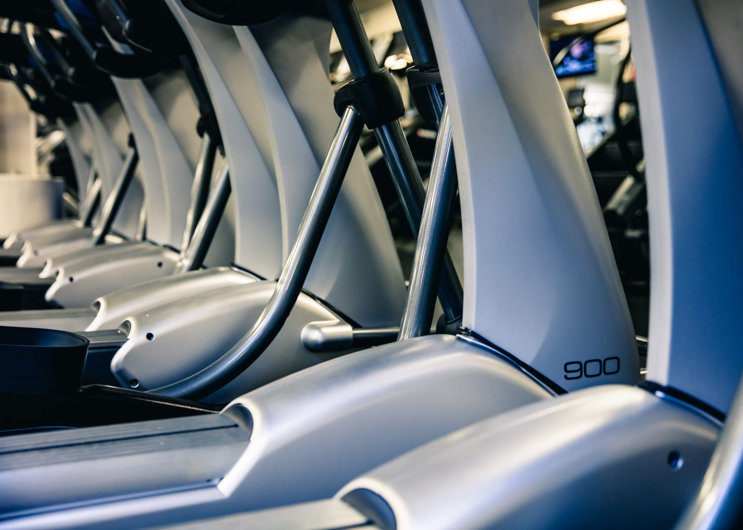 Row of elliptical machines lined up in a gym, with a focus on the metallic curved handles and control panels. The number 900 is visible on one machine in the foreground.