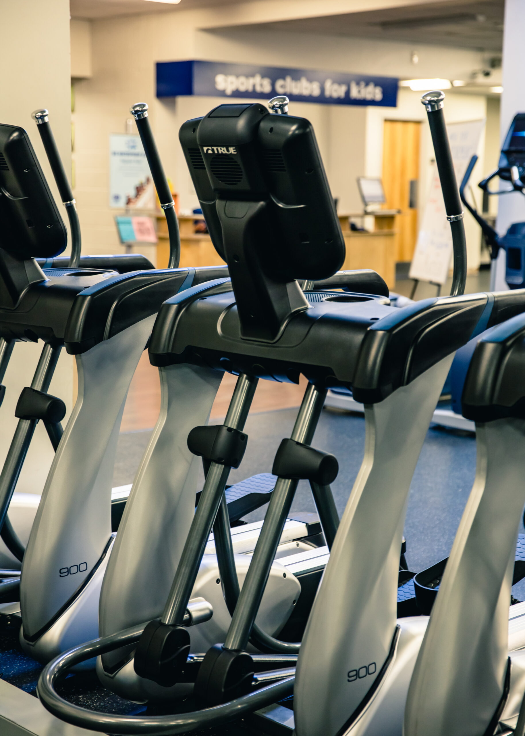 A row of elliptical machines in a gym, with digital screens and handlebars, positioned on a blue mat. A sign in the background reads sports clubs for kids.