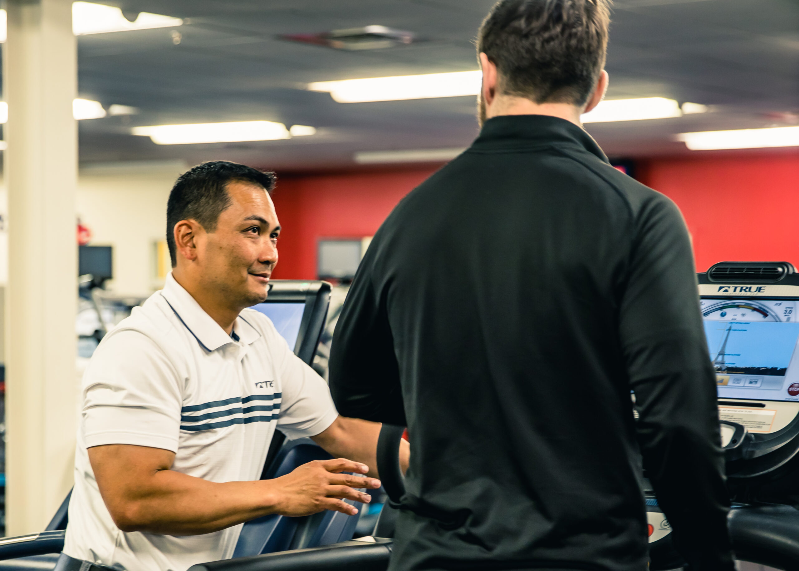 A man in a white polo shirt talks to another man in a black shirt at a gym, standing next to treadmills. The background shows gym equipment and a red wall.