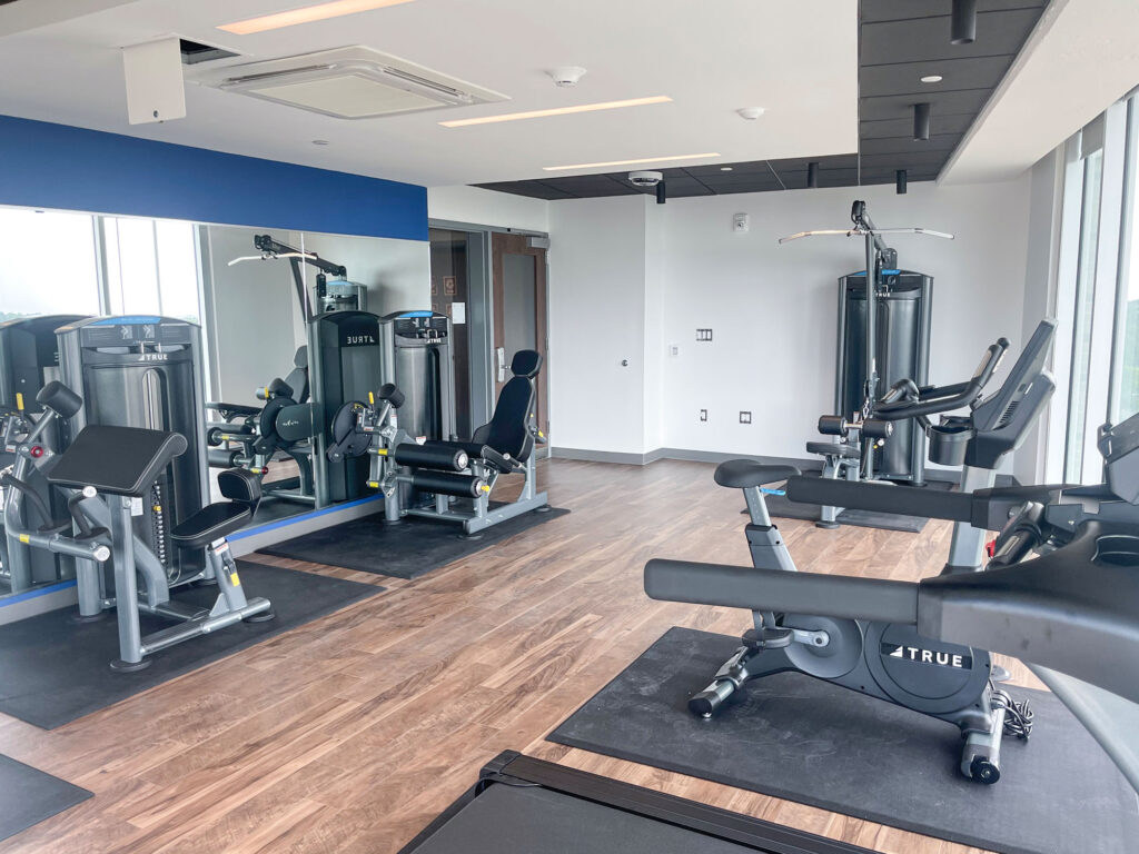 A modern gym with wood floors, large mirrors, and various exercise machines, including a leg press, lat pulldown, and stationary bike, in a bright room with large windows.