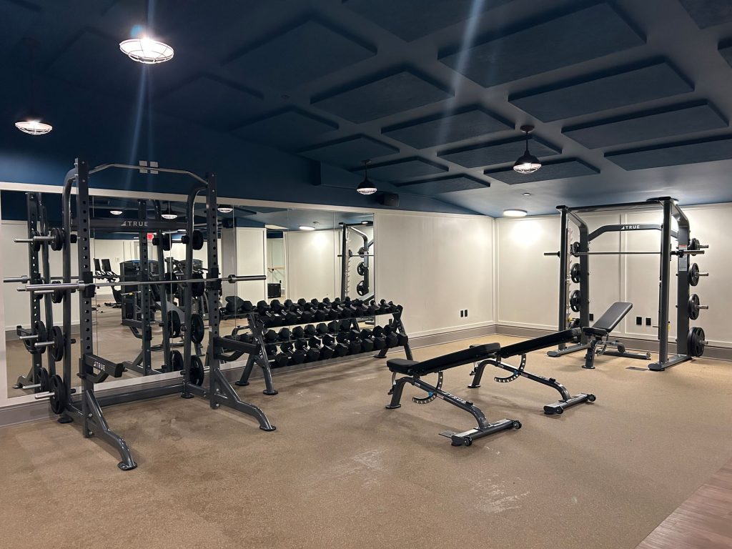 A gym with weight racks, dumbbells, benches, and two squat racks in front of mirrored walls, on a carpeted floor under ceiling lights.