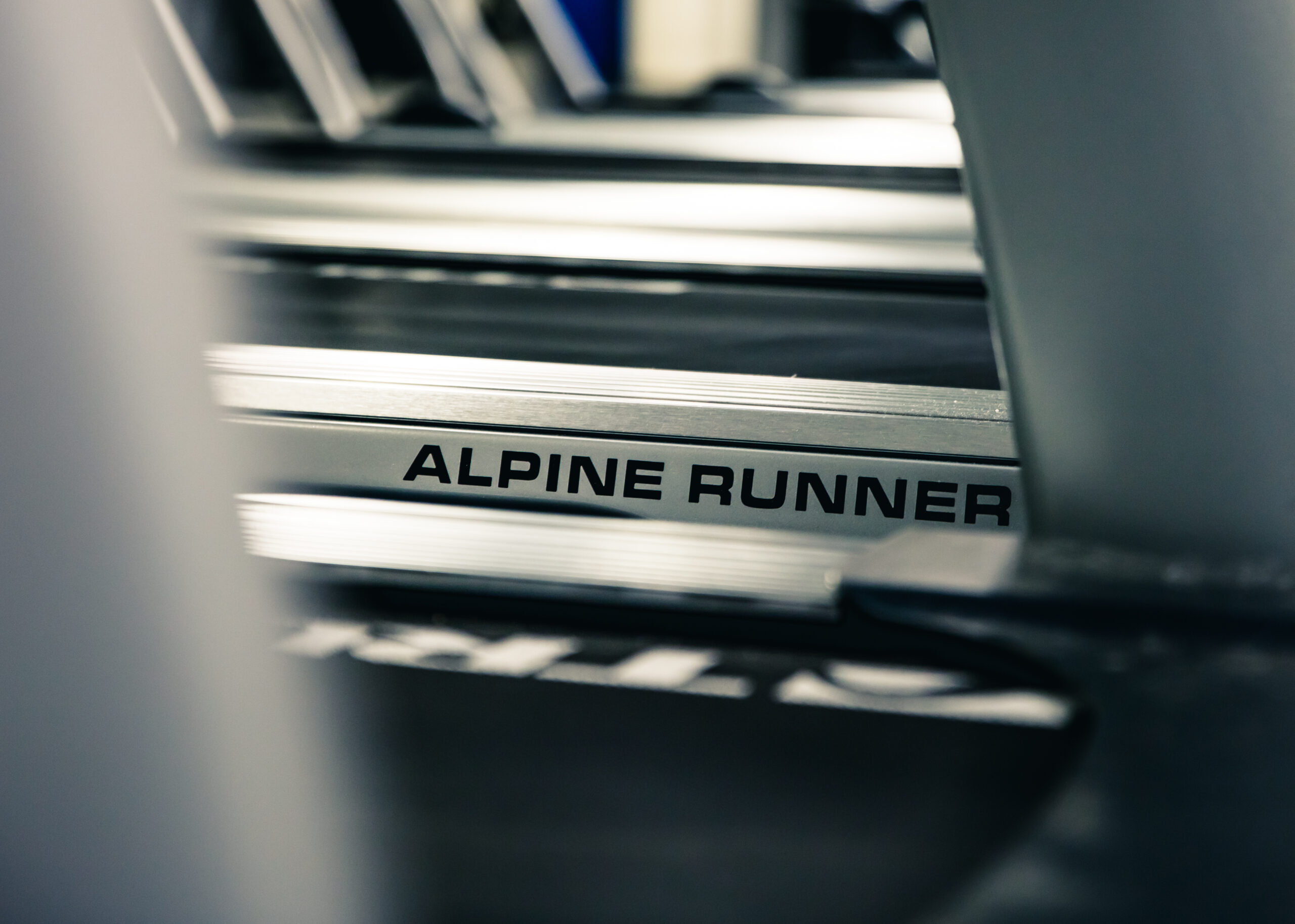 Close-up of a treadmill with the words ALPINE RUNNER printed on its side, showing part of the machine’s metal frame and blurred background gym equipment.