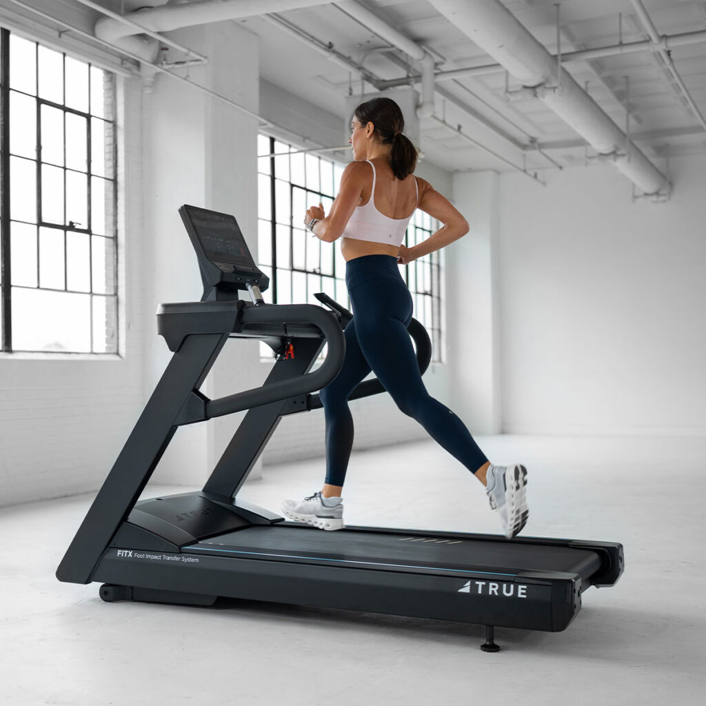 A woman in athletic wear runs on a treadmill in a bright, spacious gym with large windows and white walls.