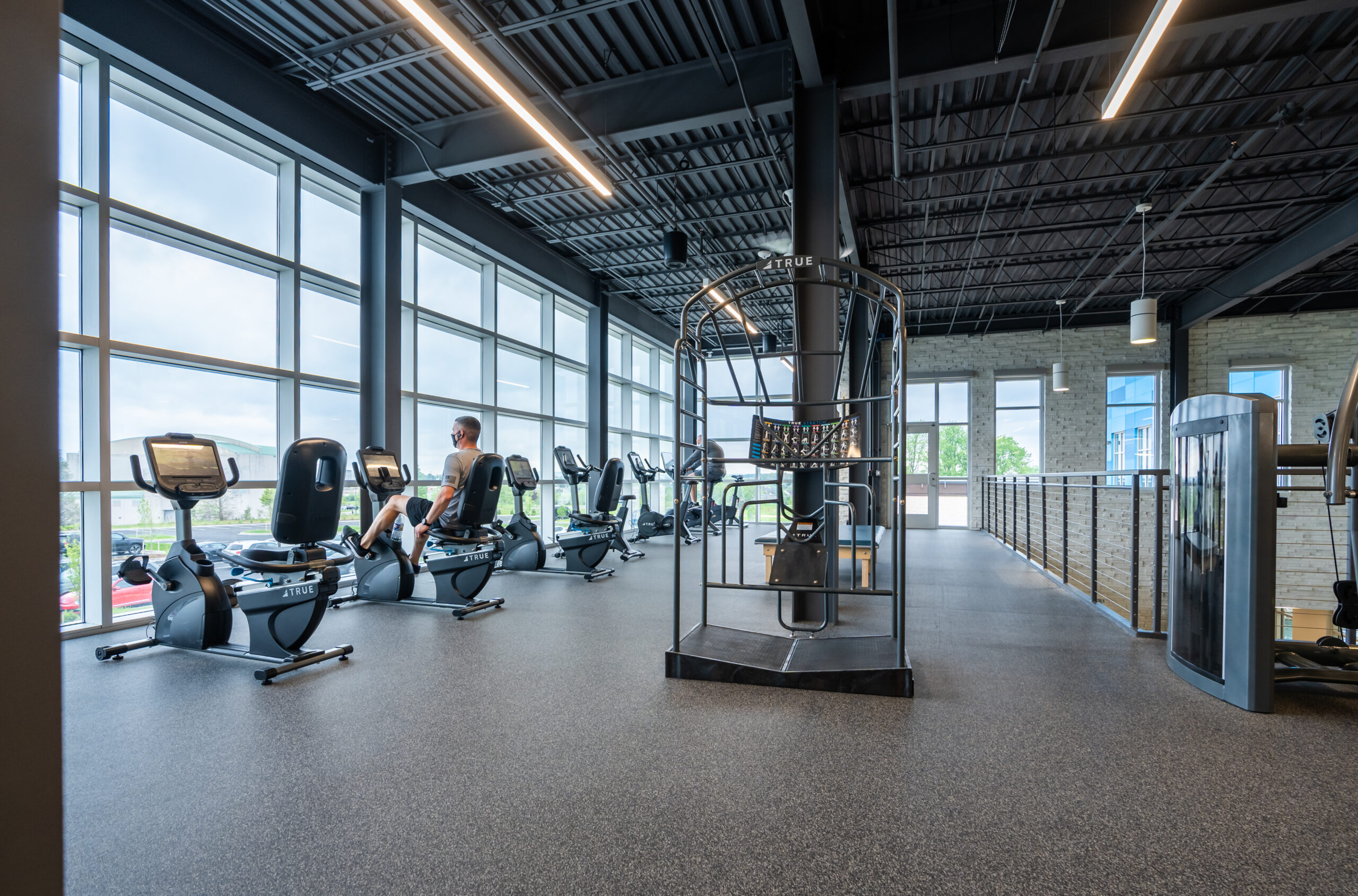 Spacious gym with large windows, exercise bikes, and a person working out. The room has modern lighting, an open ceiling, and a railing overlooking a lower level. Natural light fills the area.