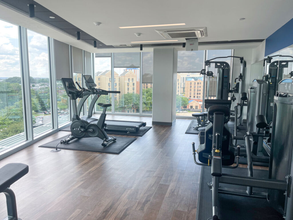 A modern gym with large windows showing a cityscape, featuring an exercise bike, weight machines, and benches on a wooden floor. Natural light fills the bright, clean workout space.
