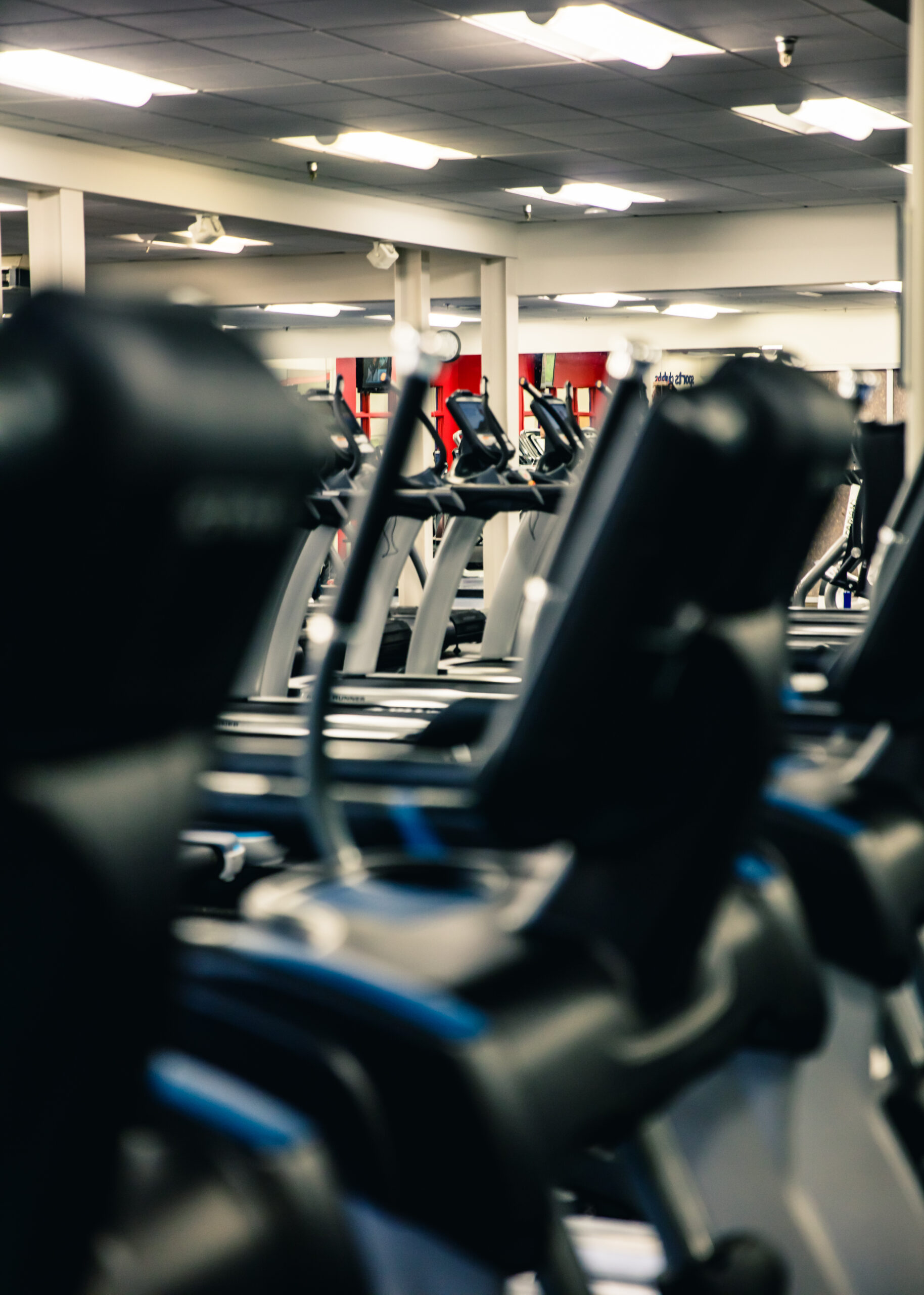Rows of elliptical machines and exercise equipment inside a brightly lit gym, with a focus on the foreground machines and mirrors reflecting the space in the background.