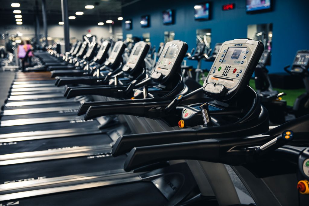 A row of empty treadmills with digital displays in a brightly lit gym, with blue walls and several TVs mounted in the background.
