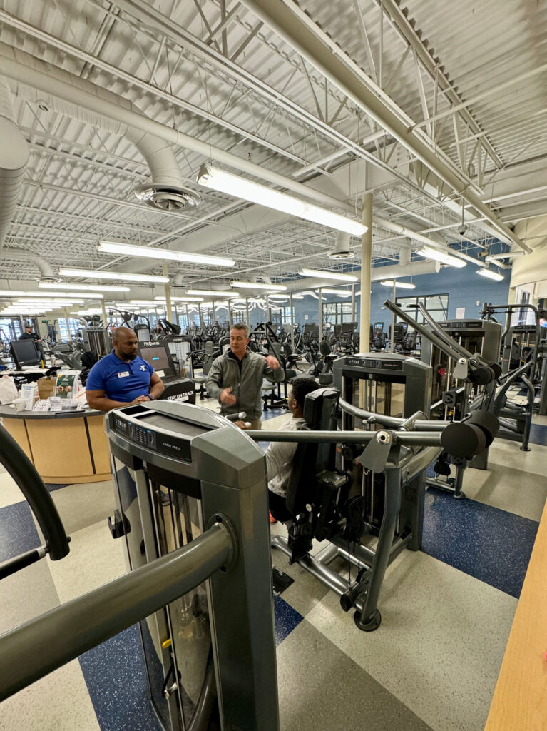 A spacious gym with several people using weight machines and cardio equipment. One person appears to be instructing another, while a third person watches. The gym has high ceilings and bright lighting.