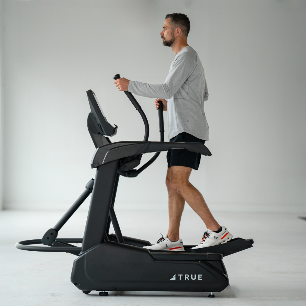 A man in a gray long-sleeve shirt and black shorts exercises on a black elliptical machine in a bright, minimalist room.