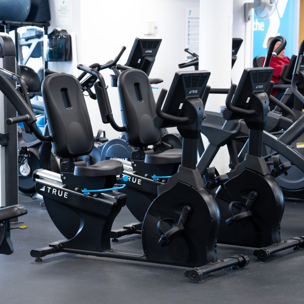 Two black recumbent exercise bikes are positioned side by side in a gym, facing forward, with various other fitness equipment visible in the background.