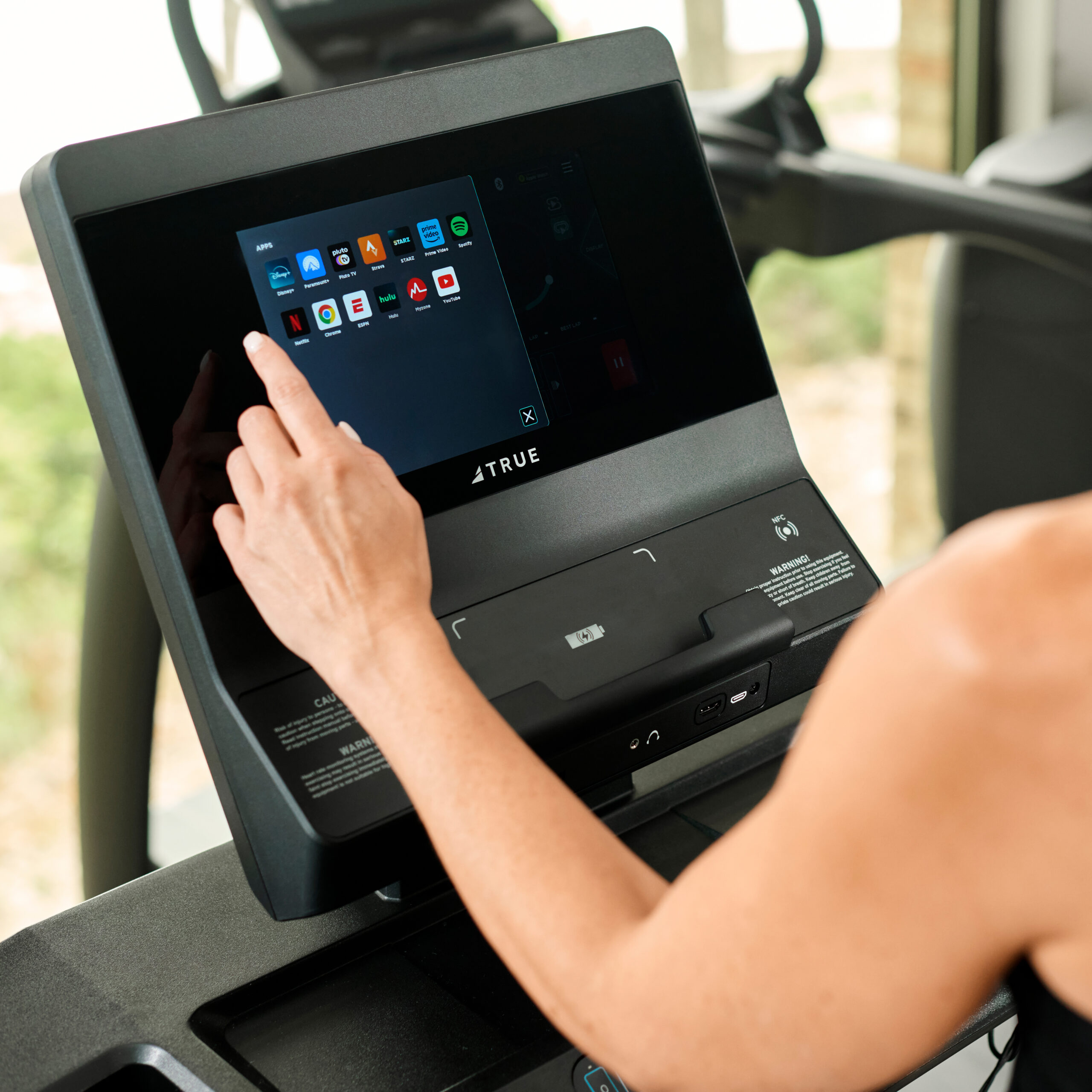 A person touches a touchscreen display on a treadmill, selecting an app from various icons. The screen is labeled TRUE and the treadmill is indoors near a window.