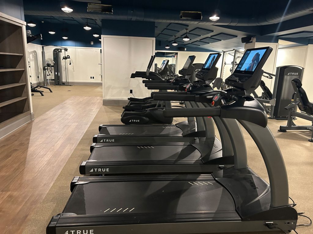 A modern gym with several treadmills in a row, each equipped with a screen, facing mirrors and other fitness equipment in a well-lit, spacious room with wooden and carpeted flooring.