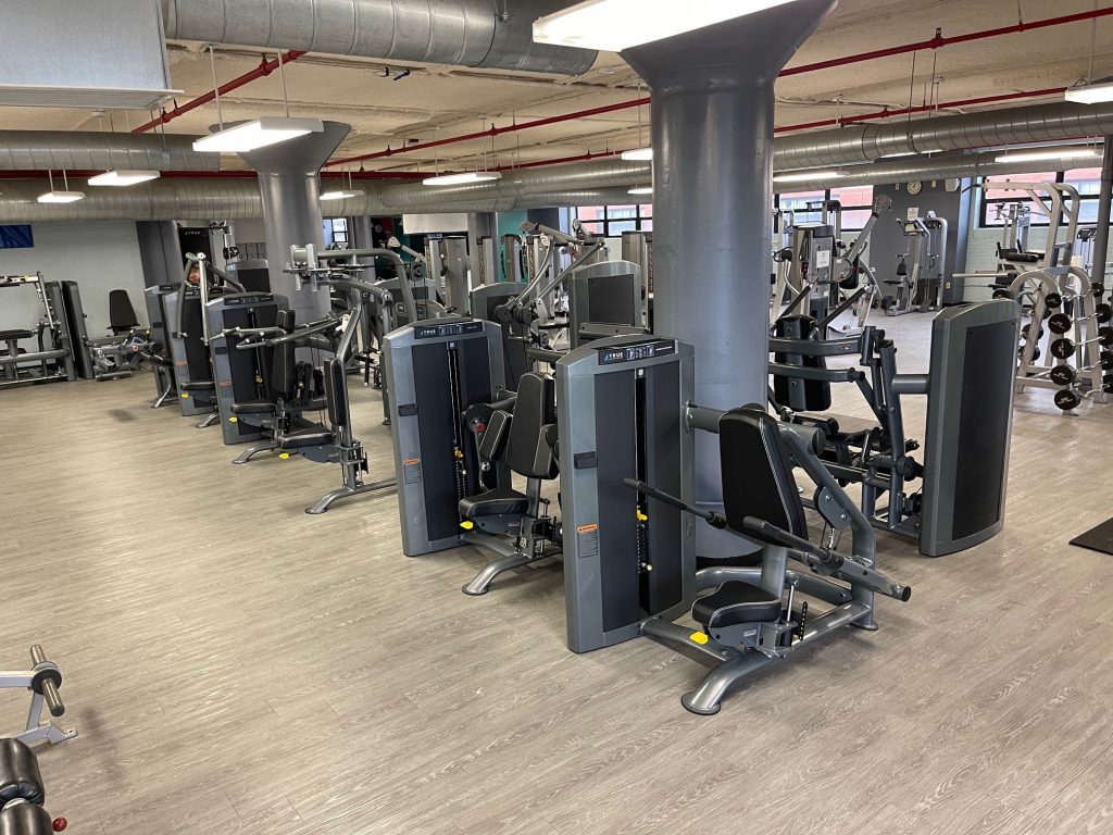 A modern gym with various exercise machines, including seated row and chest press equipment, arranged neatly on a light wood floor. The gym is spacious, with large columns and exposed ductwork on the ceiling.