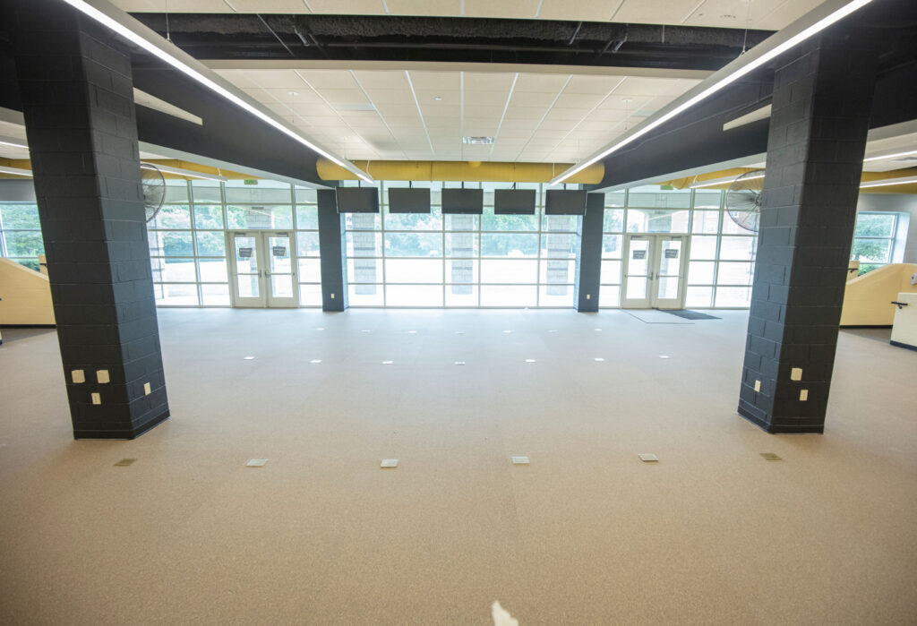 A spacious, empty room with beige floors, black columns, and large windows letting in natural light. The ceiling has fluorescent lights, and there are double glass doors at the far end.