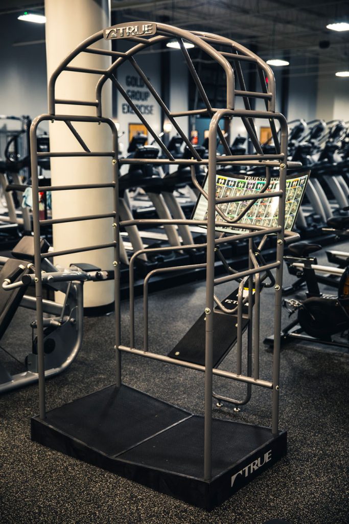 A metal workout machine with parallel bars and multiple grip positions stands on a black platform in a gym, surrounded by treadmills and other fitness equipment.
