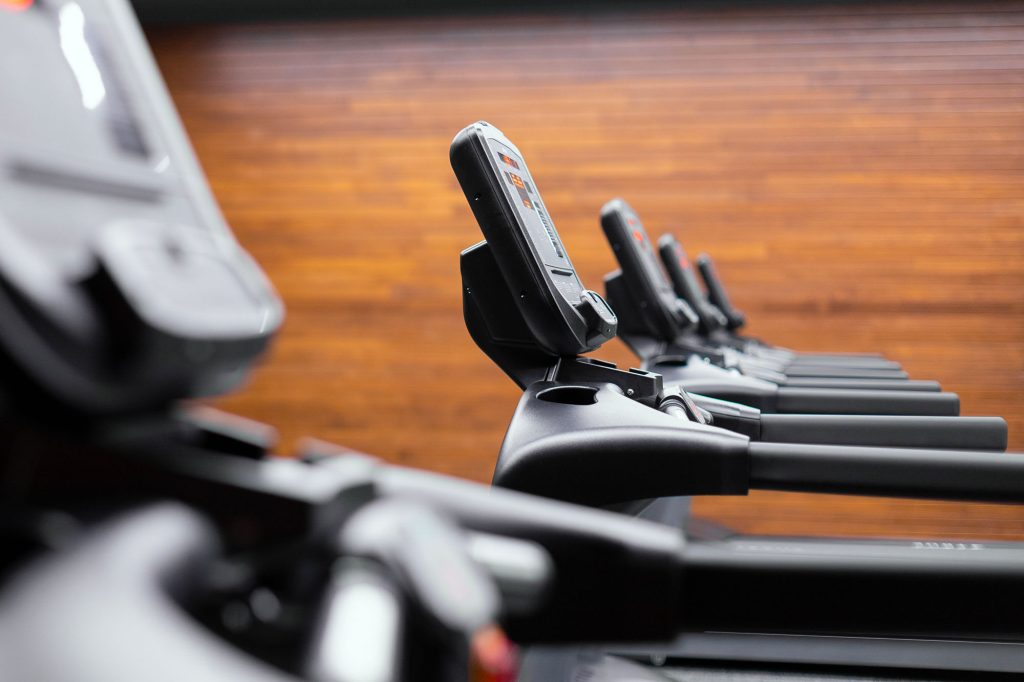 A row of modern treadmills with digital screens in a gym, with a blurred wooden wall background, viewed from the side at an angle.