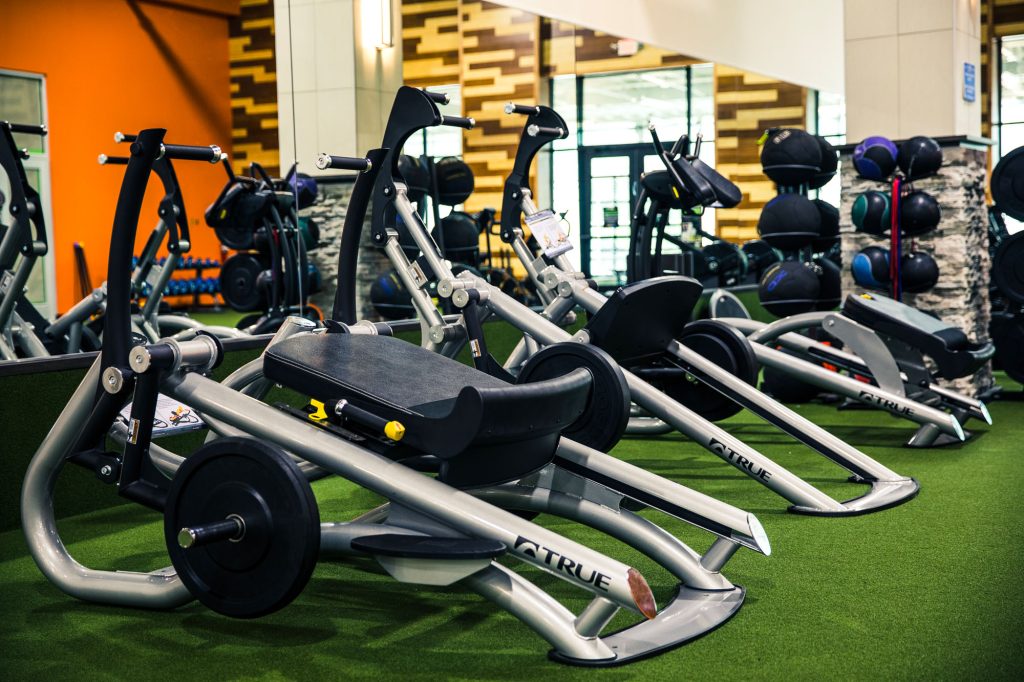 Rows of modern exercise machines in a gym, set on green turf, with various weights and equipment in the background and brightly colored walls.