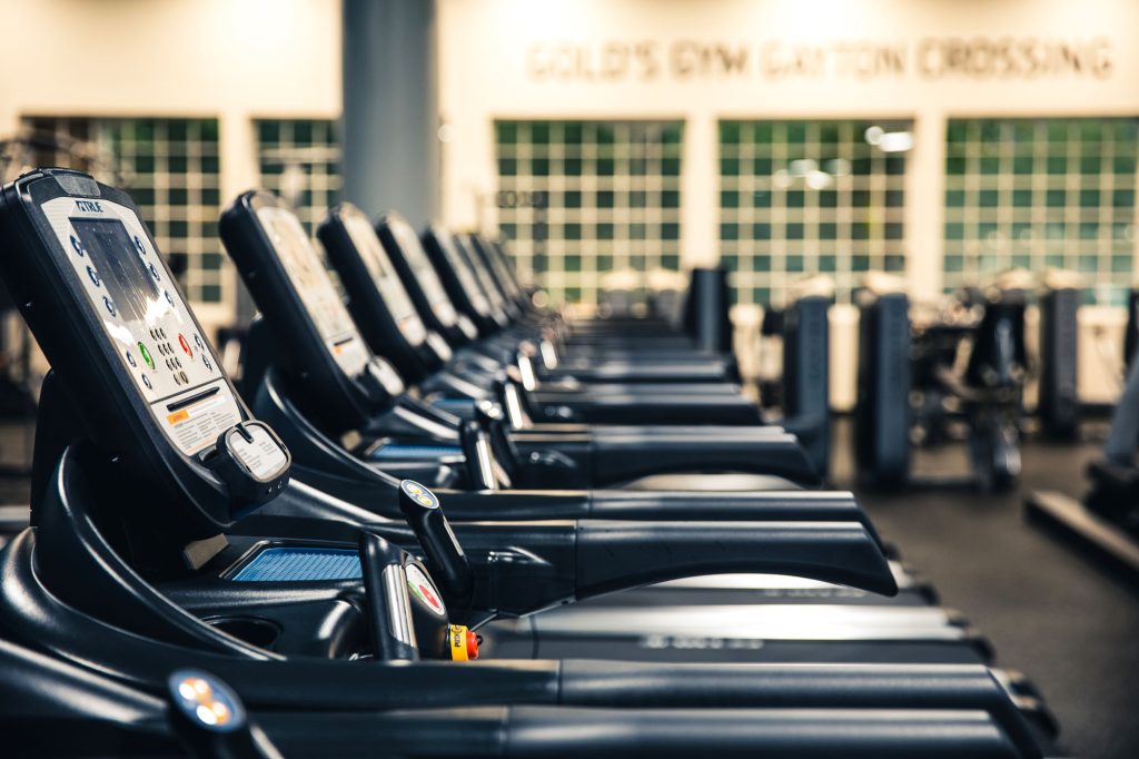 A row of treadmills in a gym, each with digital displays and control panels. The background shows blurred gym equipment and a wall with “GOLDS GYM CANTON CROSSING” written on it.